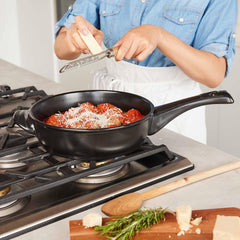 A person grating cheese over pasta in an Xtrema black ceramic skillet on a gas stove.