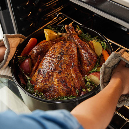 A roasted turkey with vegetables in a black Xtrema ceramic roasting pan being removed from an oven.