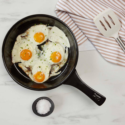 Fried eggs with herbs in a black Xtrema ceramic skillet on a white marble surface.
