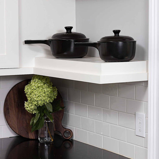 Two black Xtrema ceramic pots with lids on a white kitchen shelf above a gray tiled backsplash.