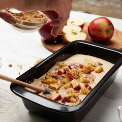Brown sugar being sprinkled over apple cake batter in a black Xtrema ceramic baking pan.