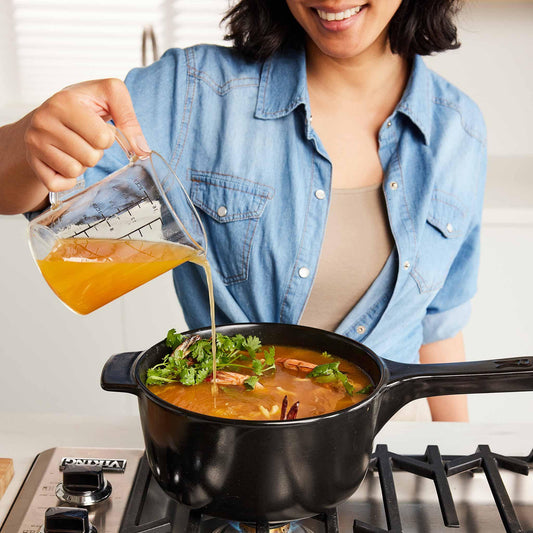 A person pouring broth into a black Xtrema ceramic pot of soup on a gas stove.