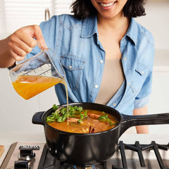 A person pouring broth into a black Xtrema ceramic pot of soup on a gas stove.