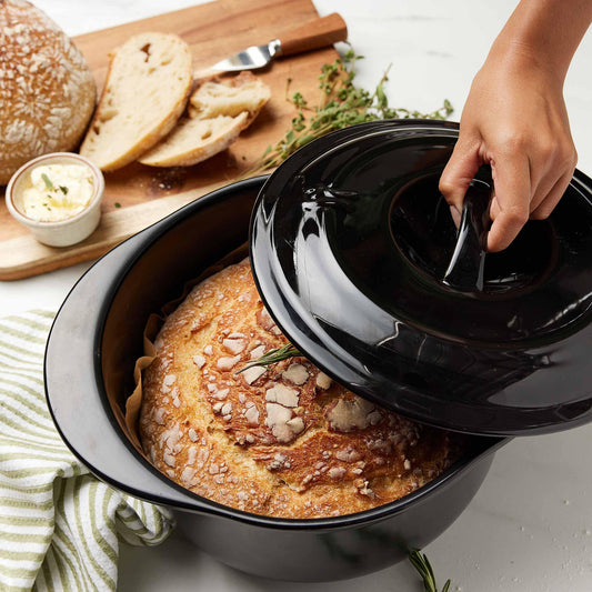 A hand lifting the black lid of an Xtrema ceramic pot to reveal a freshly baked loaf of bread.