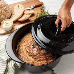 A hand lifting the black lid of an Xtrema ceramic pot to reveal a freshly baked loaf of bread.