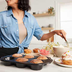 A person frosting cupcakes beside an Xtrema ceramic muffin pan filled with baked treats.