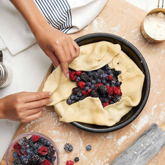 Hands folding pie crust over fresh berries in an Xtrema ceramic pie dish.
