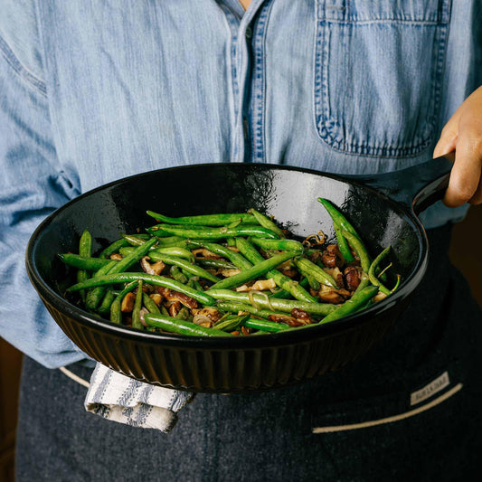A person holding an Xtrema black ceramic pan filled with sautéed green beans.