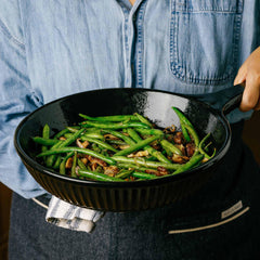 A person holding an Xtrema black ceramic pan filled with sautéed green beans.