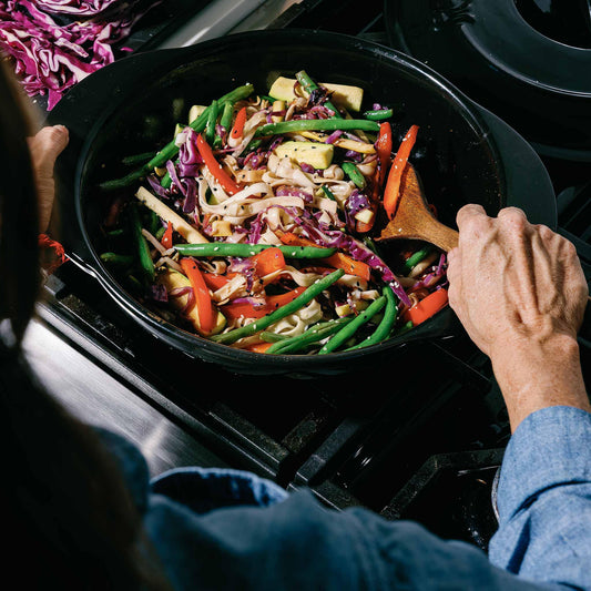A person stir-frying colorful vegetables in a black Xtrema ceramic wok.