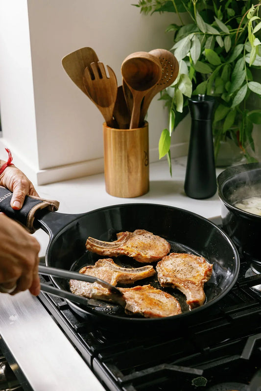 Pork chops searing in a black Xtrema ceramic skillet on a gas stove.