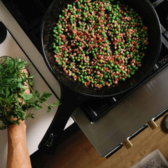 Peas and bacon cooking in a black Xtrema ceramic skillet on a stovetop.