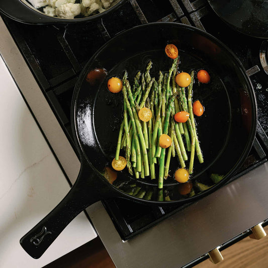 Asparagus and cherry tomatoes sautéing in a black Xtrema ceramic skillet on a stovetop.