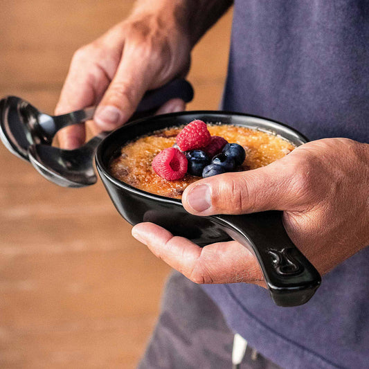 A person serving crème brûlée with berries from a black Xtrema ceramic ramekin.