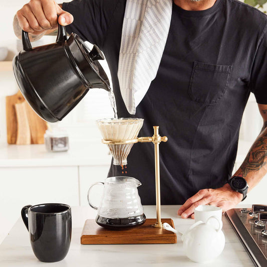 A person pouring hot water from a black Xtrema ceramic kettle into a pour-over coffee dripper.