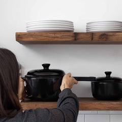 A person holding a black Xtrema ceramic pot on a wooden kitchen shelf.