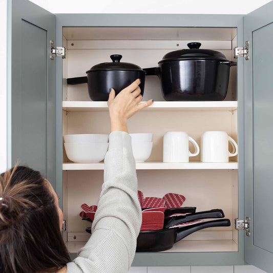 A person reaches for a black Xtrema ceramic pot inside a gray kitchen cabinet.