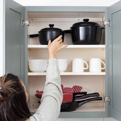 A person reaches for a black Xtrema ceramic pot inside a gray kitchen cabinet.