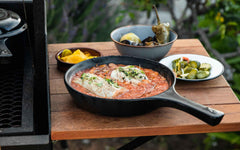 Xtrema ceramic skillet with fish in tomato sauce on a wooden table with side dishes.